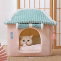 Cat peeking out from a pink and teal pet house with tassels on a light wooden floor.