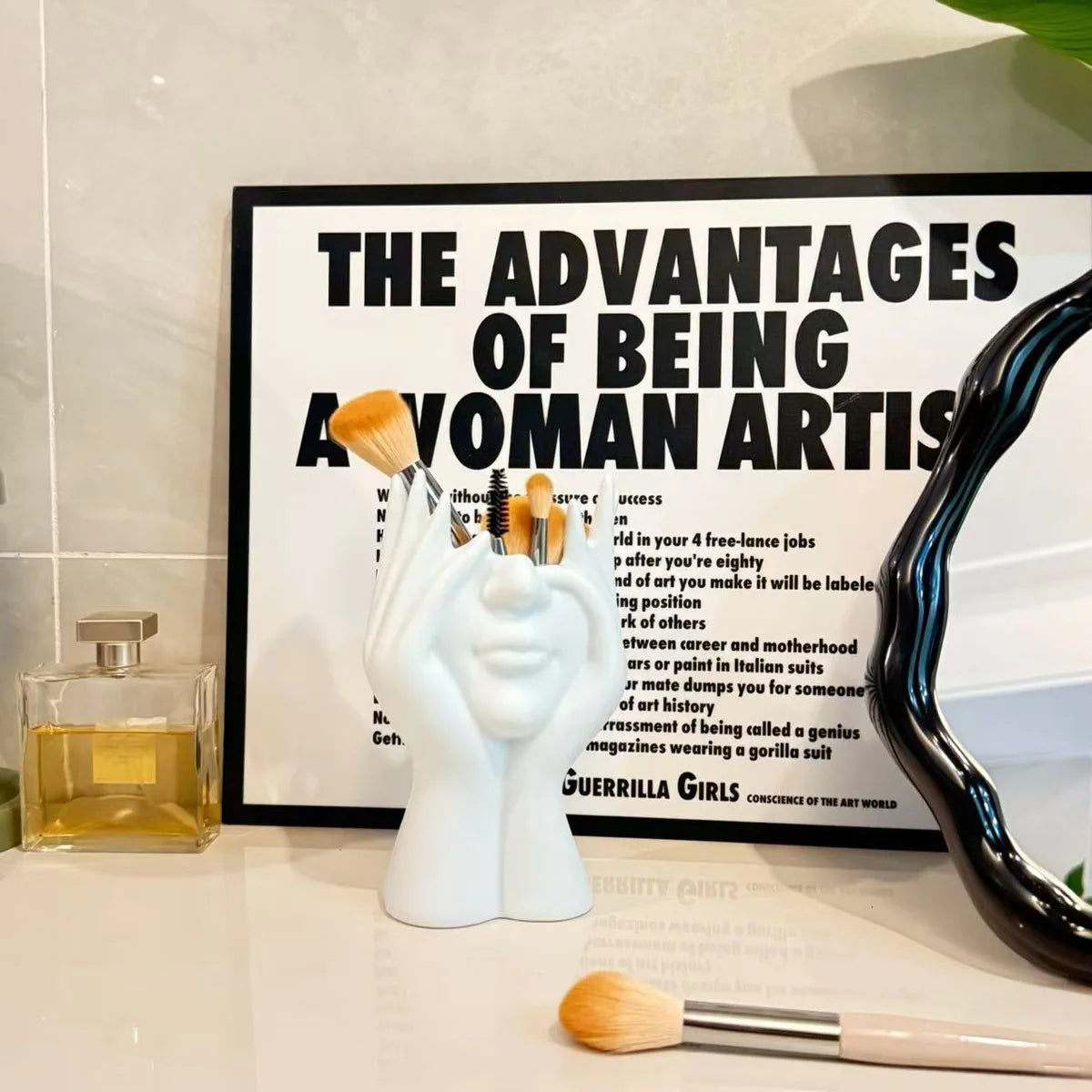 Decorative setup with a vase of brushes, a framed poster, and a mirror on a surface.