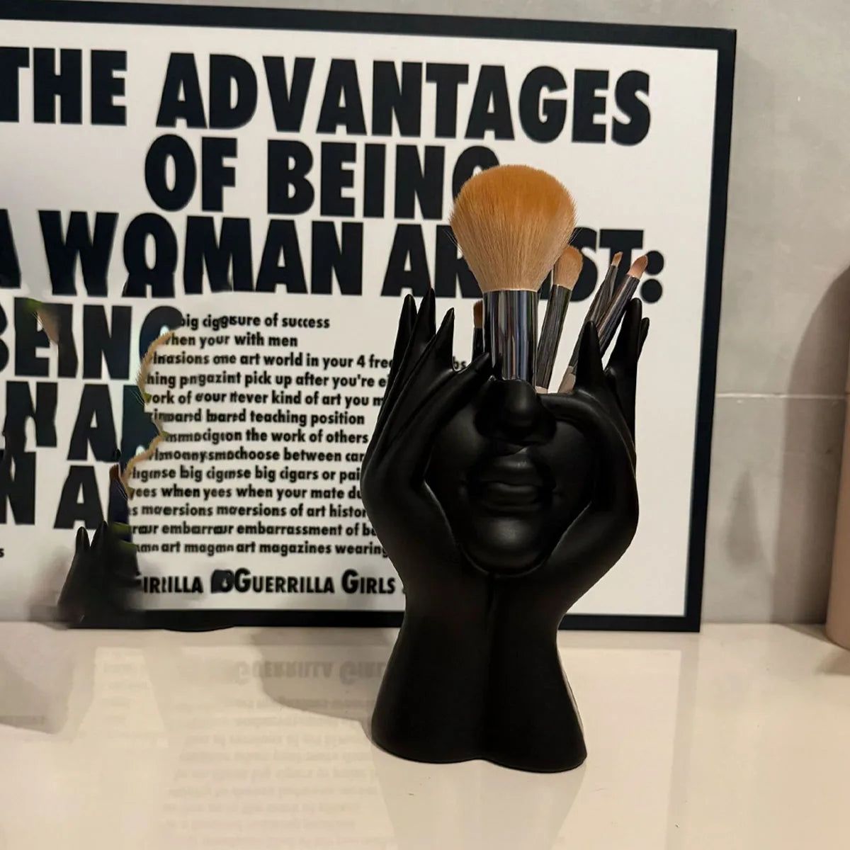Black hand-shaped brush holder with makeup brushes in front of a Guerrilla Girls art piece.