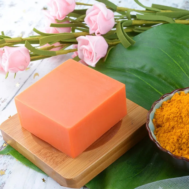 Orange bar of soap on a wooden board with pink flowers and turmeric powder in the background.