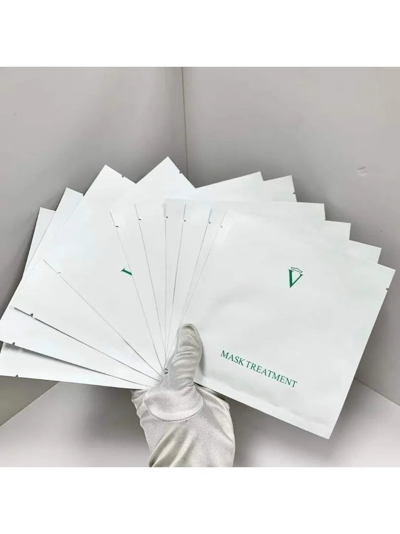 A hand wearing a white glove holds a fan of white facial mask treatment packets with green text and logo, set against a light gray background.