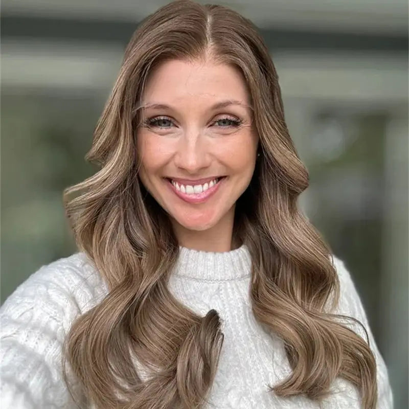 A smiling woman with long, wavy light brown hair with blonde highlights, wearing a white knit sweater, shown in a close-up portrait.