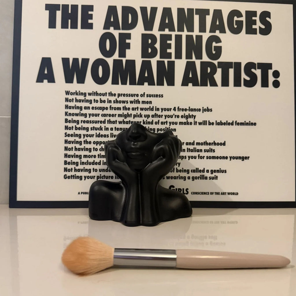Black sculpture of a woman sitting with a brush in front of a framed text piece on a white surface.