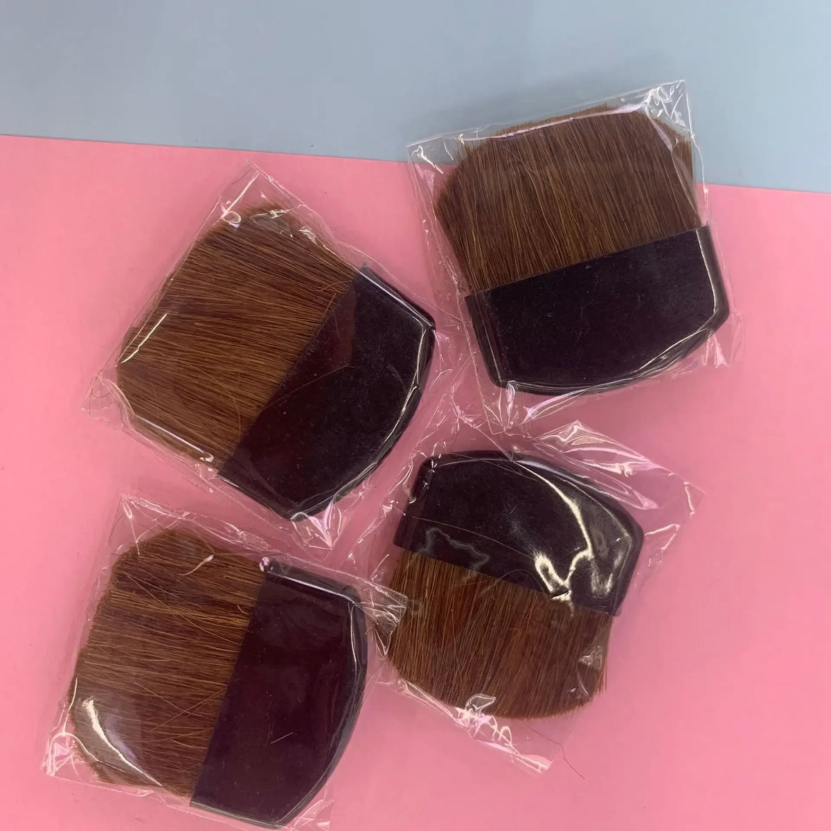 Four makeup brushes with brown bristles in clear packaging on a pink surface.