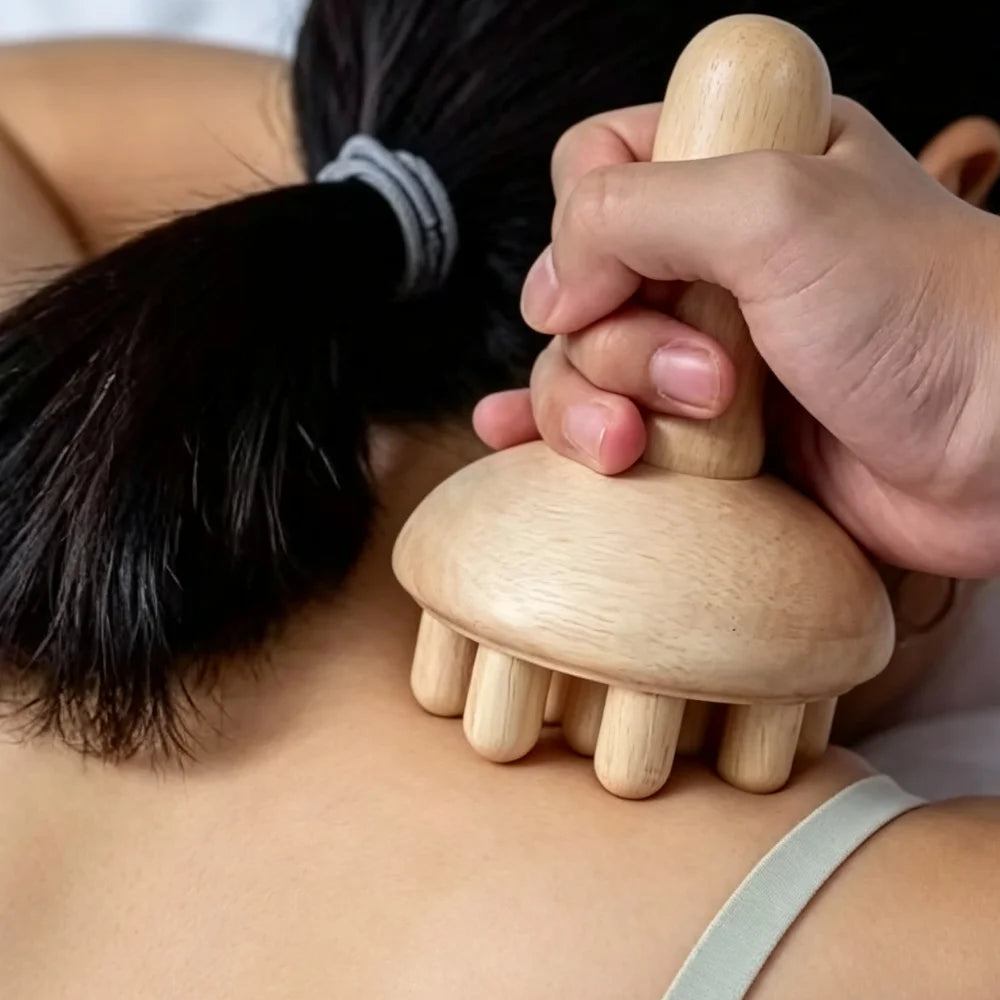 A close-up of a person using a wooden scalp massager on their upper back, with the hand holding the handle and the massager head resting on the skin.