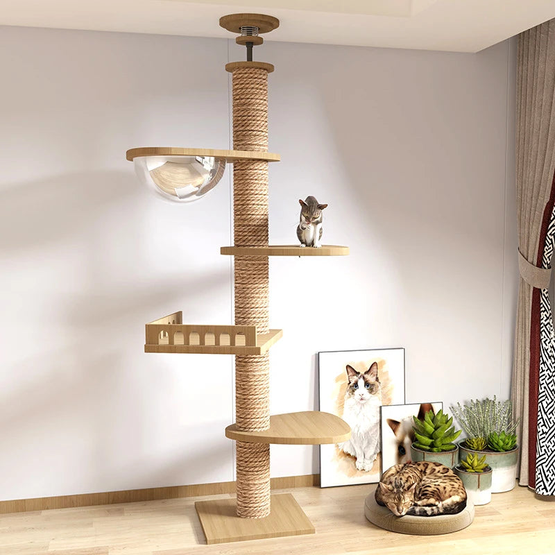 A tall wooden cat tree with a rope-wrapped post, multiple perches, a clear glass bowl, and a scratching post section, shown in a styled room with two cats on and around it. The scene includes framed cat art, small potted plants, and curtains.