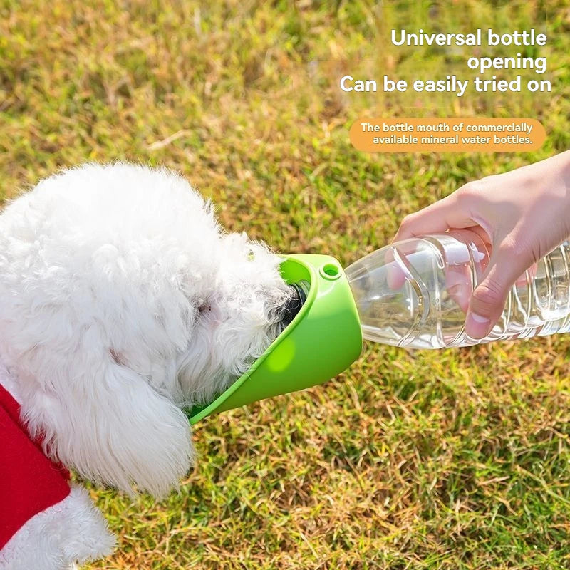 A white dog wearing a red collar drinks water from a clear plastic bottle using a bright green universal adapter. The scene is set outdoors on a grassy lawn, with a human hand holding the bottle.