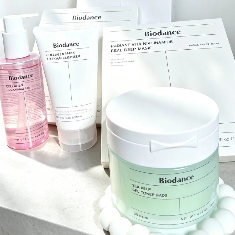 Biodance skincare set featuring a Sea Kelp Gel Toner Pads jar in the foreground, a pink Collagen Cleansing Gel bottle, and a white To Foam Cleanser tube, with their respective white boxes behind them on a light marble surface.