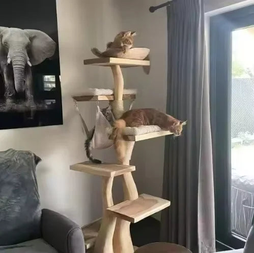 A tall wooden multi-level cat tree with cushioned platforms and a hanging hammock, placed in a living room next to a window with gray curtains. Two cats are resting on the cat tree.