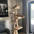 A tall wooden multi-level cat tree with cushioned platforms and a hanging hammock, placed in a living room next to a window with gray curtains. Two cats are resting on the cat tree.