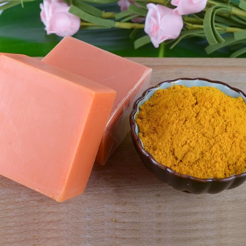 Orange soap bar next to a bowl of yellow powder on a wooden surface with pink flowers in the background.