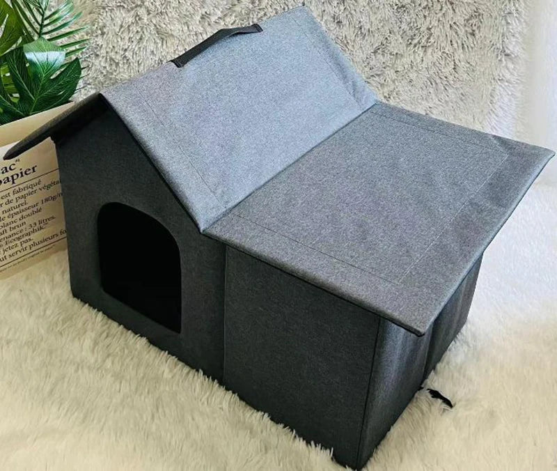 A gray fabric pet house with a slanted roof and arched entryway, placed on a white faux-fur surface with a plant and book visible in the background.