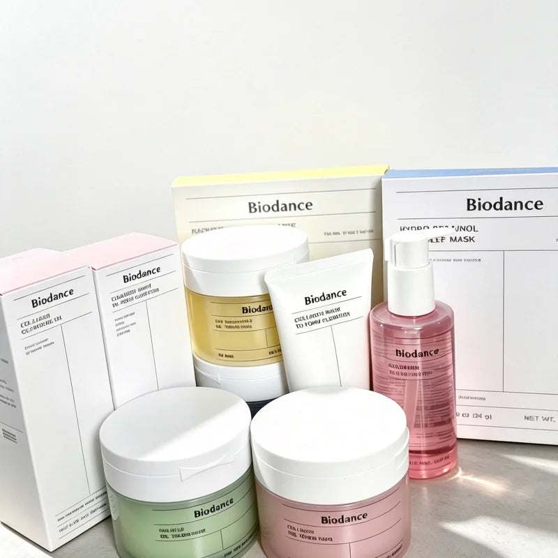 A set of Biodance skincare products arranged on a light surface against a white background, including jars, tubes, and boxes in white, pale pink, pale yellow, green, and pink.