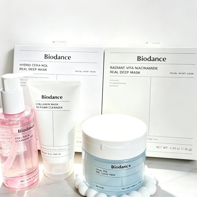 Biodance skincare set featuring a pink bottle of facial essence, a white tube of collagen mask to foam cleanser, and a light blue jar of coral hibiscus clay mask, arranged in front of their white product boxes on a clean white background.