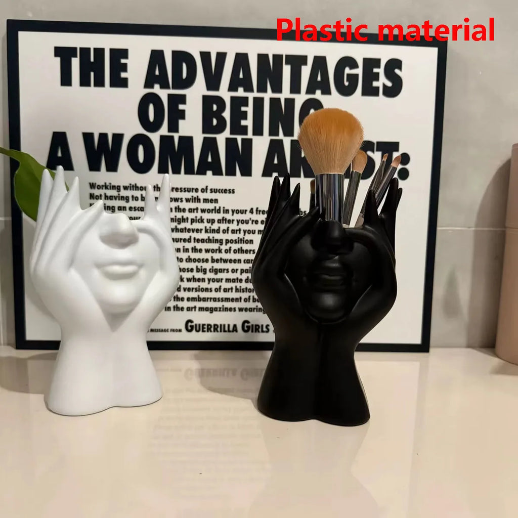 Black hand-shaped brush holder with makeup brushes on a white surface, with a white wall and framed text in the background.