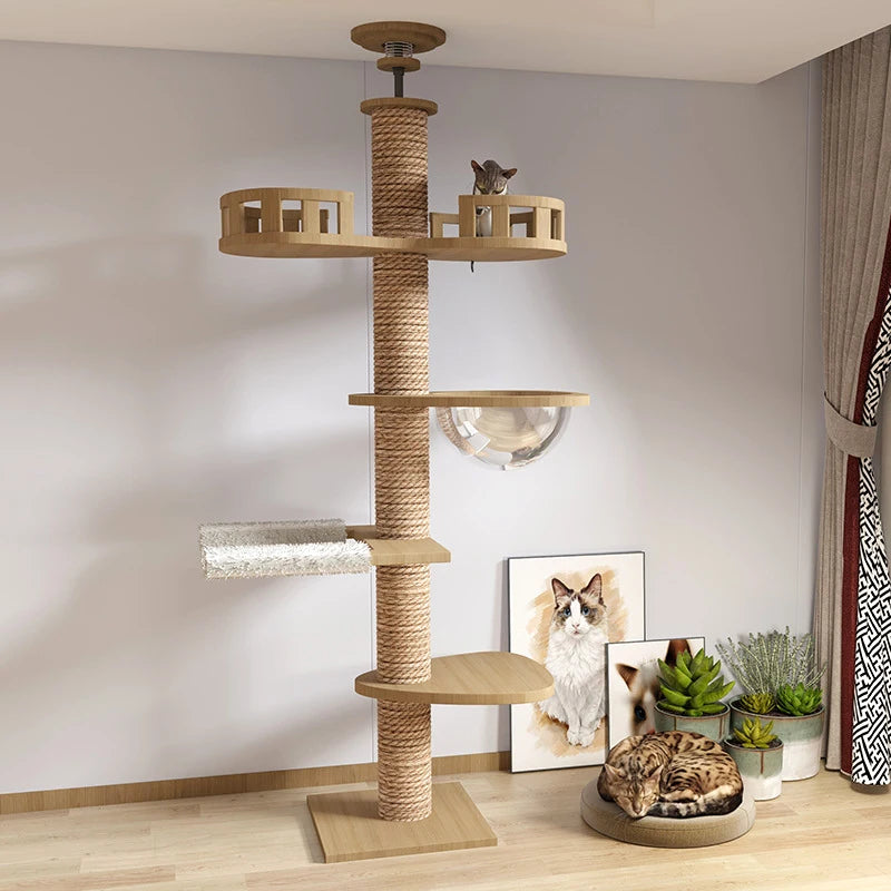 A tall wooden cat tree with rope-wrapped posts, multiple platforms, a clear hanging bowl, and a top perch, shown in a styled room with a cat on the top platform and another cat sleeping on a bed below. A framed cat photo and small potted plants sit on the floor against a light wall.