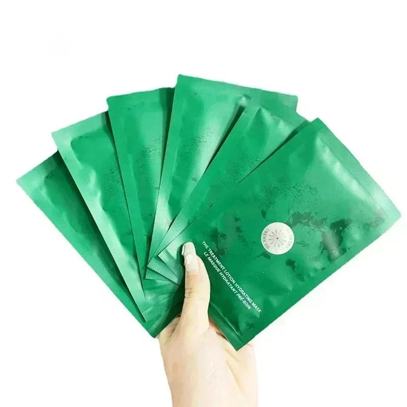 A hand holding a fanned-out set of five shiny green skincare mask packets against a plain white background.