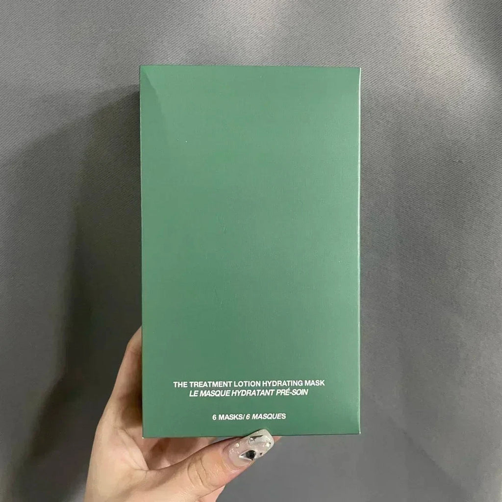 A hand with decorated fingernails holds a green rectangular box of 6 Masks 6 Masks Hydrating Treatment Lotion against a gray fabric background.