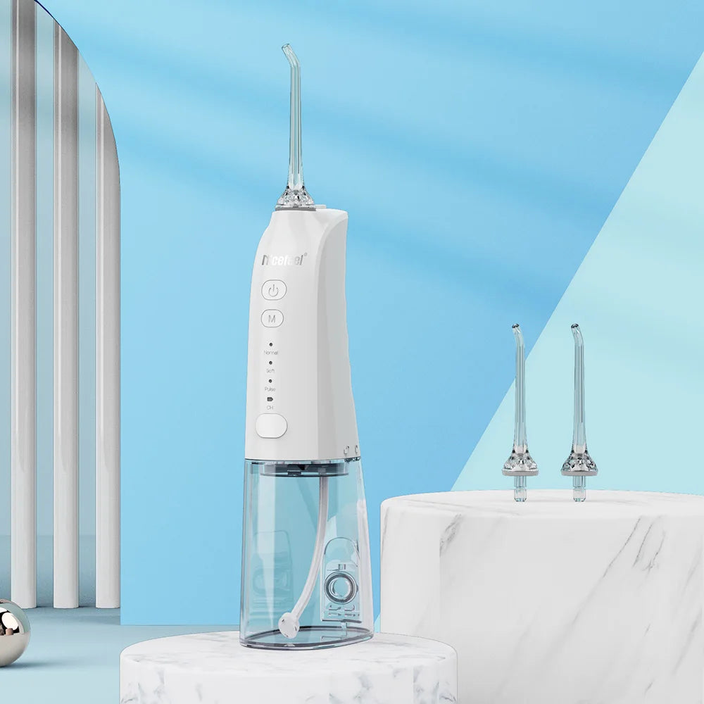 A white and clear electric oral irrigator water flosser stands on a white marble pedestal against a blue and light blue studio background. Two additional nozzle tips are displayed on a marble sink-like surface in the background.