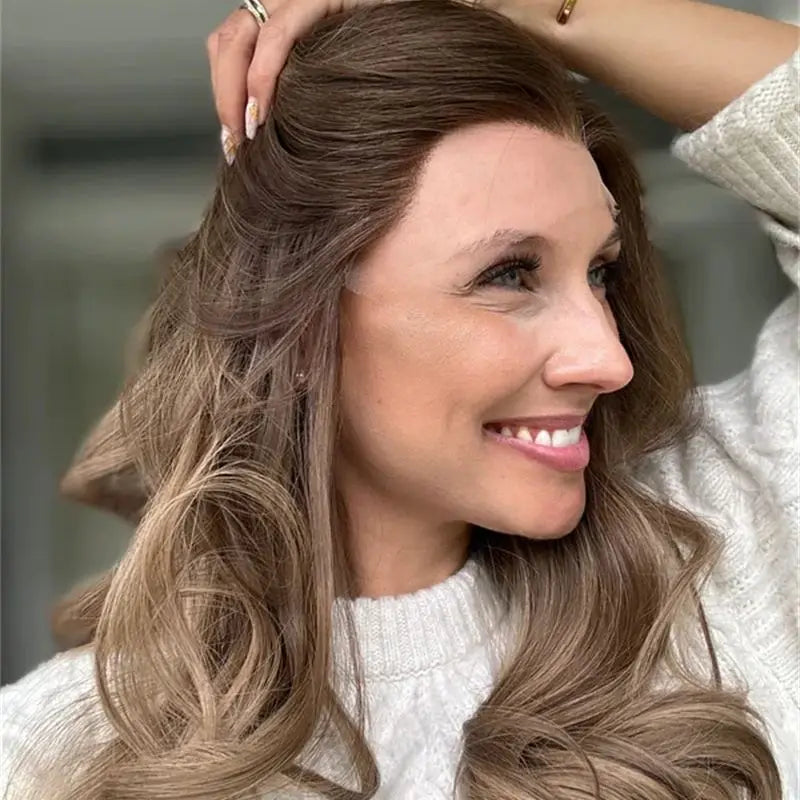 A woman with long, wavy brown hair with lighter blonde highlights, wearing a white knit sweater and smiling while looking to the side.