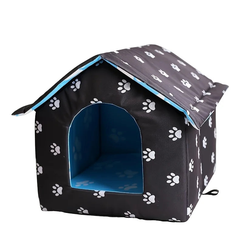 A small house-shaped pet bed with a black exterior covered in white paw prints, a blue interior, and a matching blue underside, shown on a plain white background.