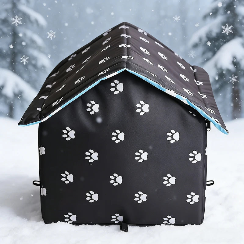 A small house-shaped dog bed covered in a black waterproof fabric with a white paw print pattern, set in a snowy outdoor scene with blurred evergreen trees and falling snow.