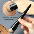 Makeup brush with black handle and wooden ferrule, held by a hand, with foundation cream visible.