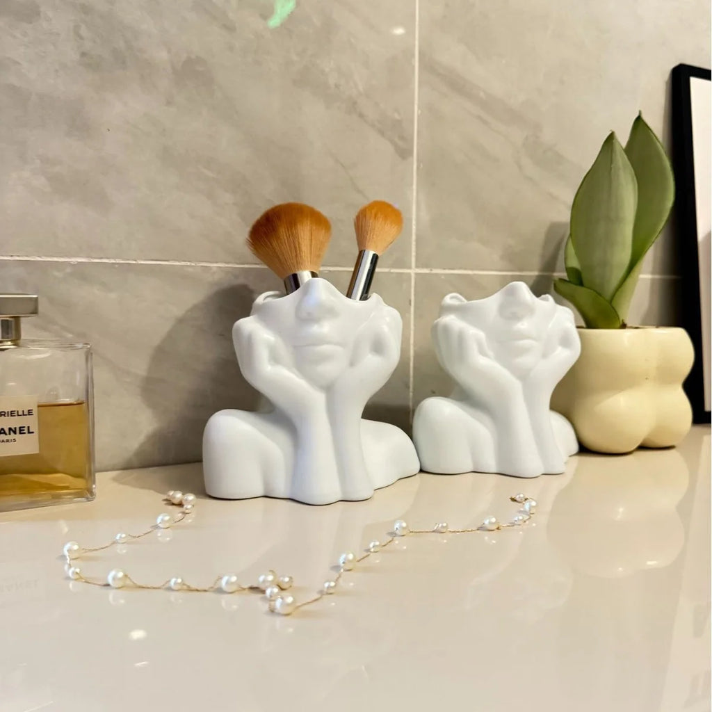 Two white ceramic face-shaped holders with makeup brushes on a bathroom counter.