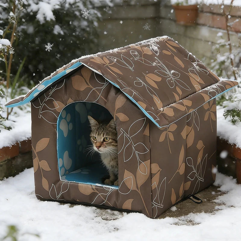 A small house-shaped cat bed with a brown and beige leaf pattern and blue interior, set outdoors in snow with a tabby cat peeking out from the entrance.