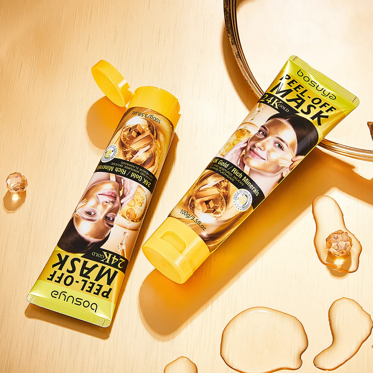 Two tubes of bosuya Peel-Off Mask in a yellow and gold color scheme, arranged on a warm beige wooden surface with decorative liquid droplets and a mirror edge.