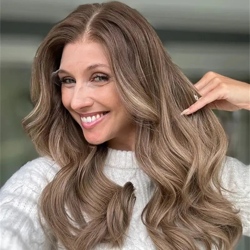 A smiling woman with long, wavy brown hair with blonde highlights, wearing a white knit sweater. A hand is gently holding a section of her hair to show texture and color.