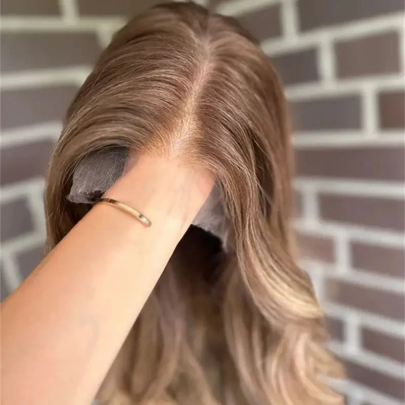 A close-up of a person's hand holding a lace front wig against a gray brick wall background. The wig has a medium brown root with blonde highlights throughout the length.