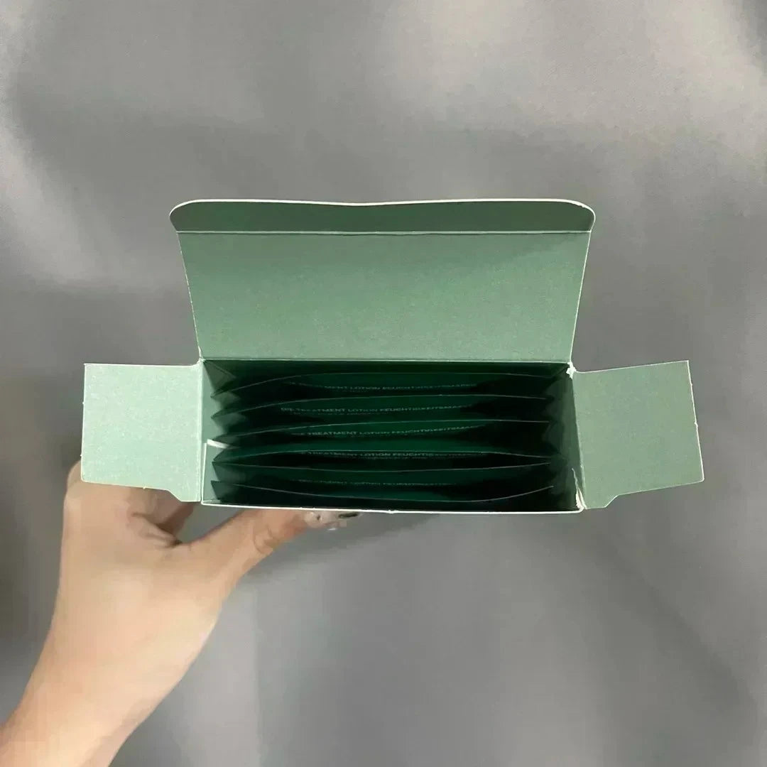 A hand holding an open sage green box containing multiple dark green sachets, photographed against a plain gray background.