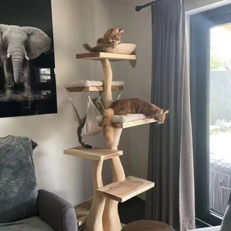 A tall wooden multi-level cat tree with cushioned platforms and a hanging hammock, placed in a living room next to a window with gray curtains. Two cats are resting on the cat tree.