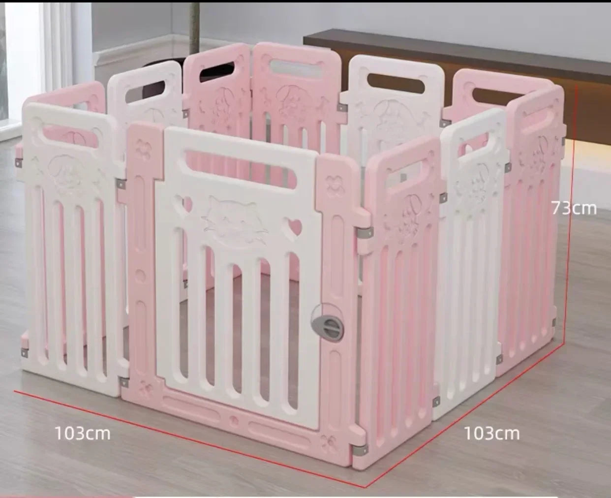 Pink and white plastic pet playpen with a gate, shown on a light wood floor with overlaid dimension lines and text indicating 103cm by 103cm by 73cm measurements.