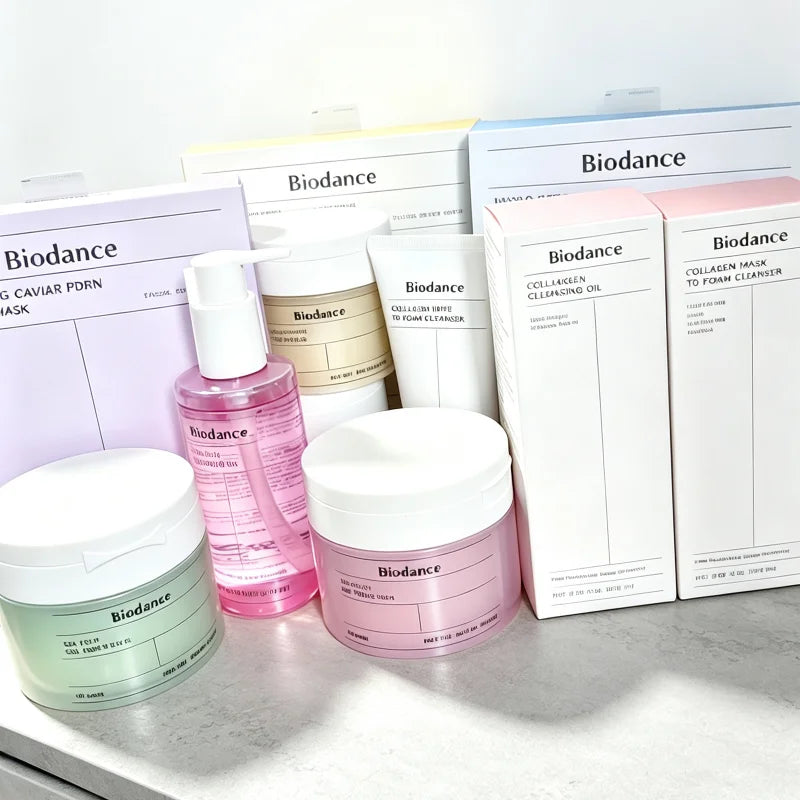 A Biodance skincare set arranged on a light surface, including multiple white jars, a pink pump bottle, and pastel-colored product boxes with visible Biodance branding.