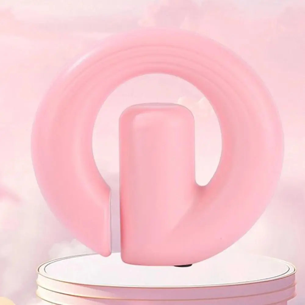 A pink silicone ring-shaped product with a central cylindrical protrusion, displayed on a white round pedestal against a soft pink background.