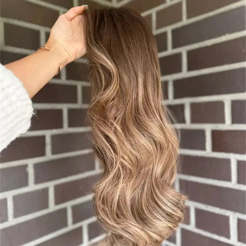 A long, wavy wig with a brown-to-blonde ombré color, held up by a hand against a dark brick wall background.