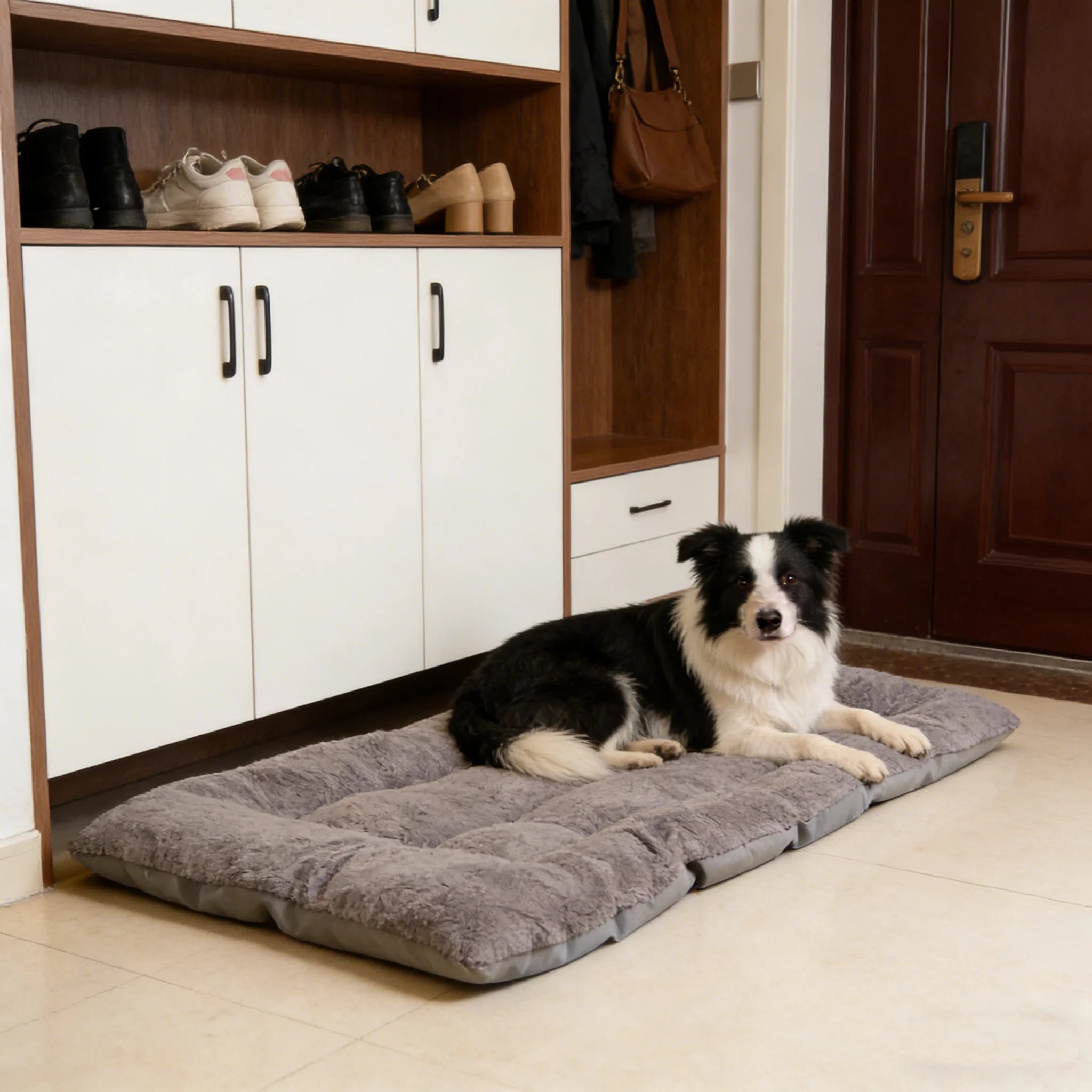 A black and white dog lies on a gray plush entryway mat on a tiled floor in front of white and wood entryway cabinets.